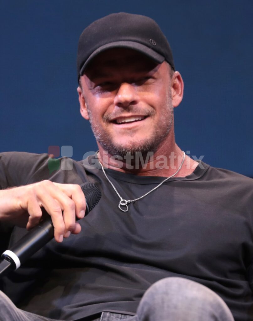 Alan Ritchson smiling and holding a microphone at a 2023 event in Phoenix, Arizona